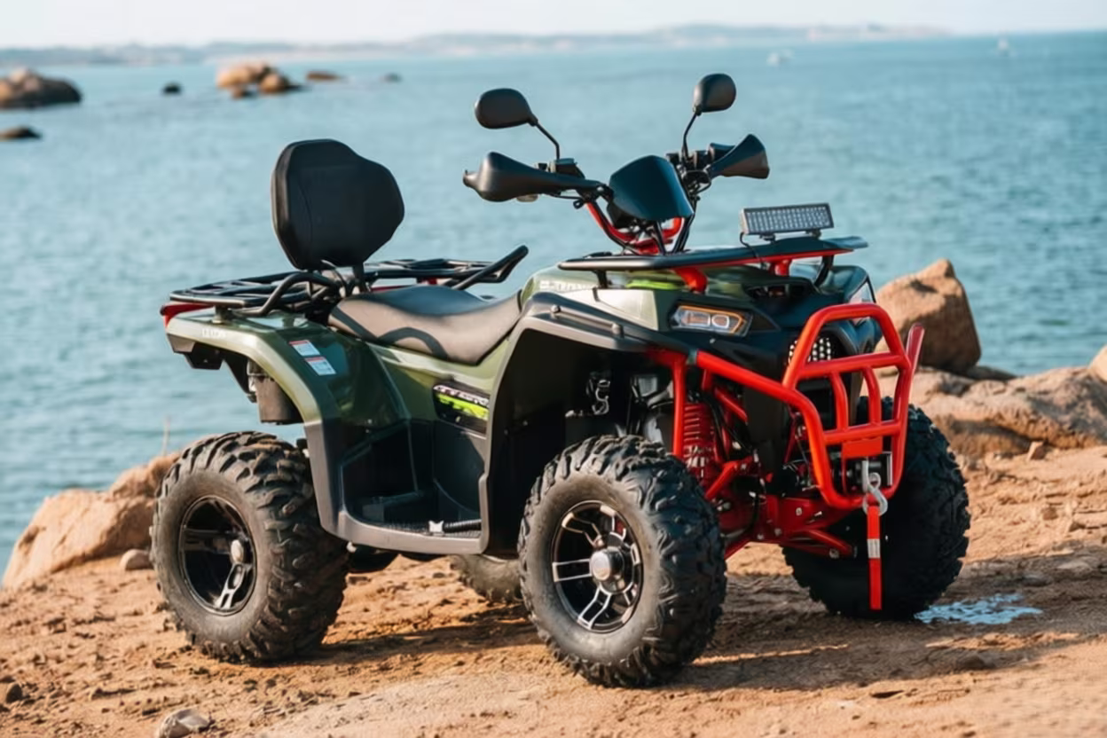 A red-framed ATV with black and green body parked on a sandy shore near the ocean, featuring rugged tires, a rear cargo rack, and a front bumper, set against a scenic coastal backdrop.
