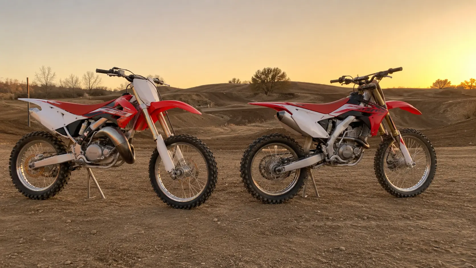 Two dirt bikes parked on a track, with a slim 2-stroke on the left and a thicker 4-stroke on the right.